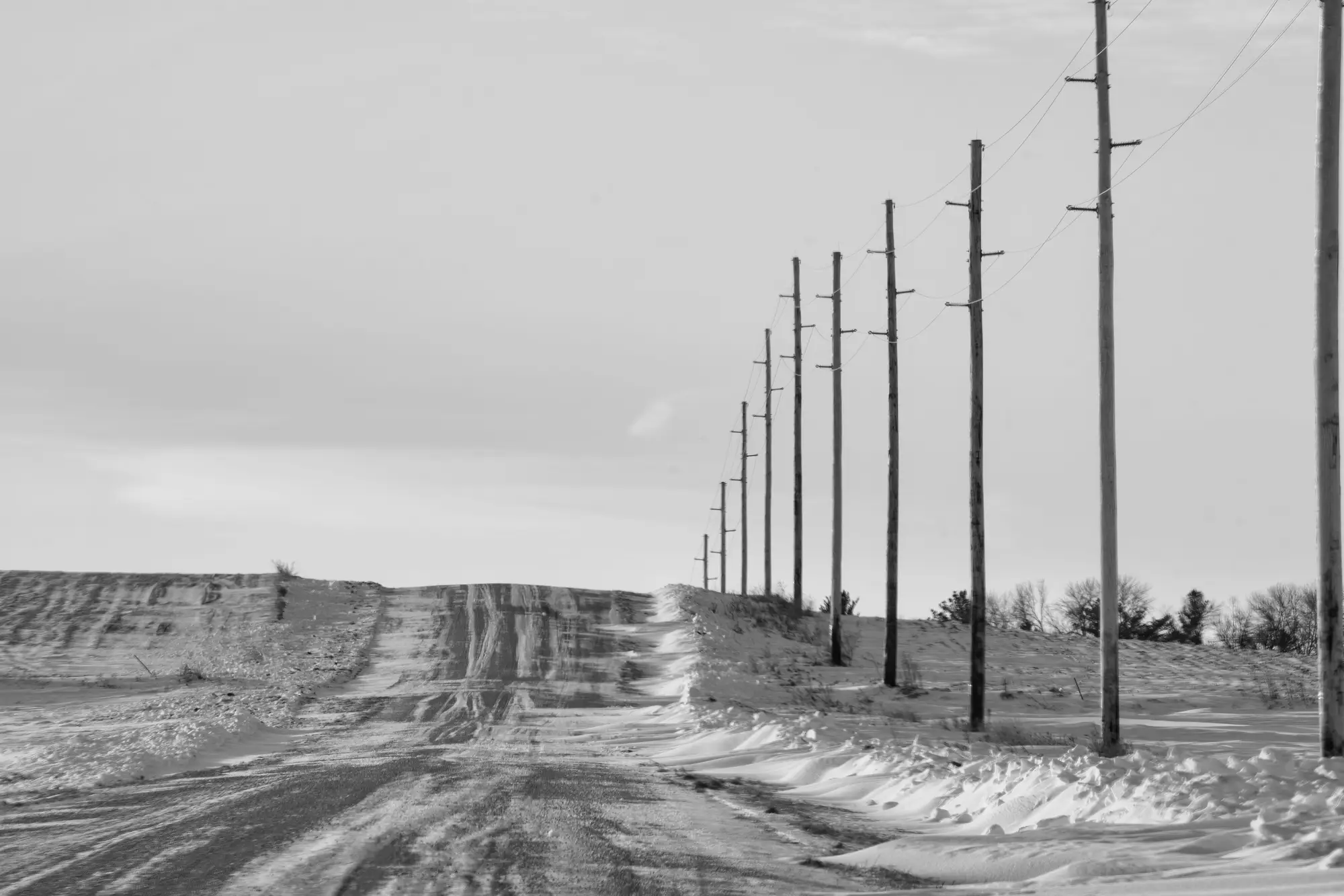Wooden distribution poles with crossarms running beside a snowy county road, in black and white
